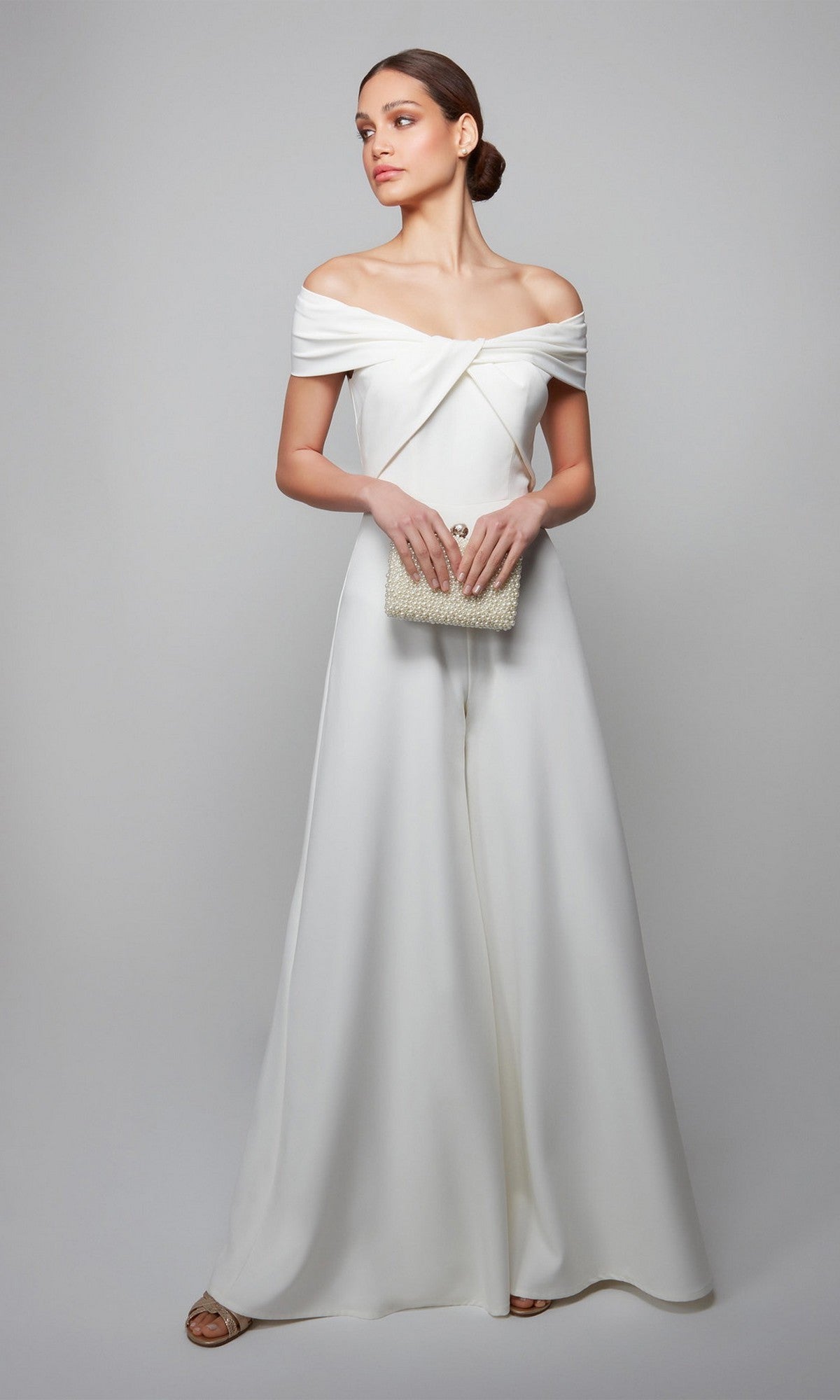 A woman wearing the Long Formal Jumpsuit: Alyce 70013 in off-the-shoulder white holds a textured beige clutch while standing against a plain light gray background.